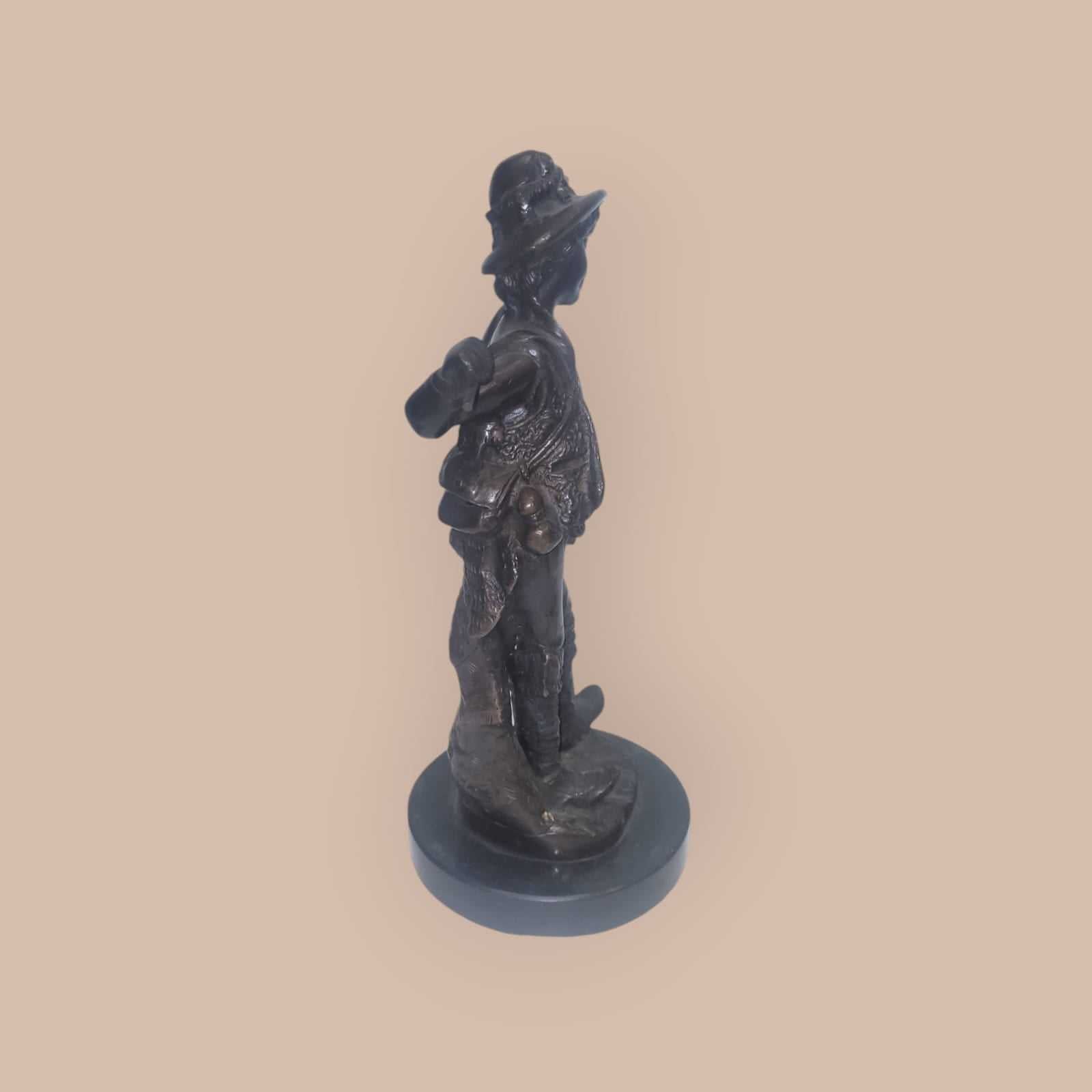 Escultura de bronce antigua de niño con gorro y bastón, vista lateral que resalta su textura y diseño ornamental para decoración de interiores.