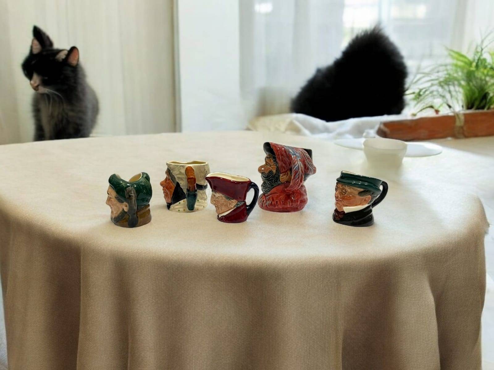 Jarras Royal Doulton de personajes famosos sobre mesa redonda con mantel beige, decoración clásica para hogar con estilo inglés.