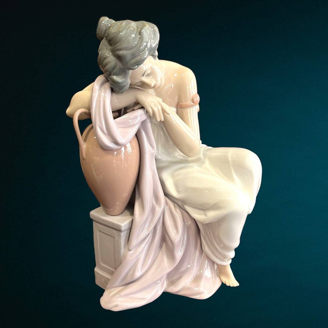 Escultura decorativa Lladró 'Soñando Despierta' con mujer en pose melancólica sobre ánfora y pedestal, elaborada en porcelana artesanal.