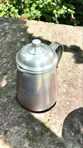Jarra de aluminio vintage de 1/2 litro con tapadera colocada en superficie de piedra al aire libre. Aporta un toque nostálgico a espacios exteriores.