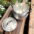 Olla de Aluminio Vintage abierta con tapa al lado, colocada sobre troncos en jardín. Perfecta para decoración de estilo retro en exteriores.