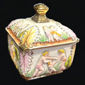 Bombonera de porcelana decorada con detalles dorados y escenas clásicas en relieve. Ideal para decorar y almacenar dulces. Medidas 17x17xH21 cm.