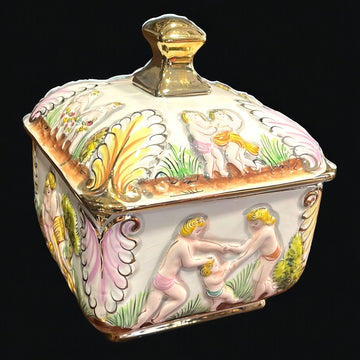Bombonera de porcelana decorada con detalles dorados y escenas clásicas en relieve. Ideal para decorar y almacenar dulces. Medidas 17x17xH21 cm.