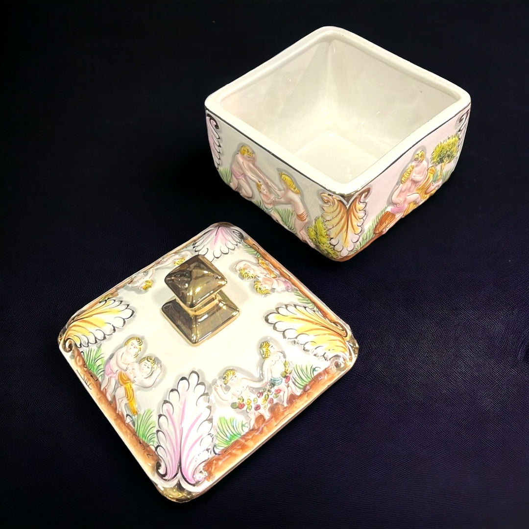 Bombonera de porcelana con tapa removible, decorada con relieves de figuras clásicas y detalles dorados. Perfecta para embellecer cualquier espacio.