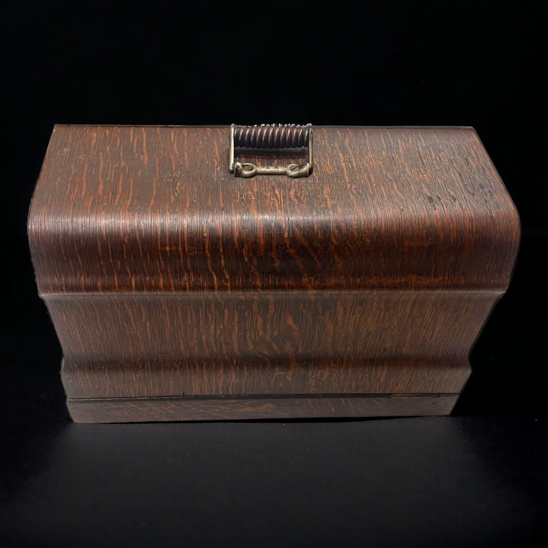 Vista frontal de la caja de madera con acabado envejecido y asa metálica, utilizada para almacenar la máquina de coser portátil New Ideal.