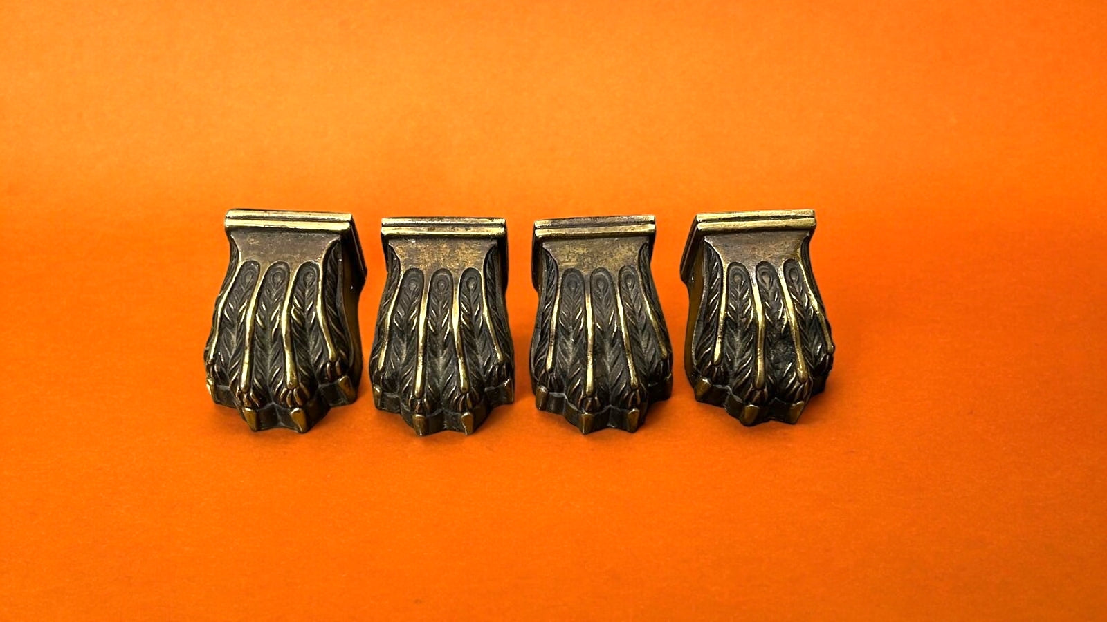 Cuatro garras de bronce para patas de mesa, con acabado detallado y sofisticado, sobre fondo naranja. Perfectas para decoración clásica y elegante.
