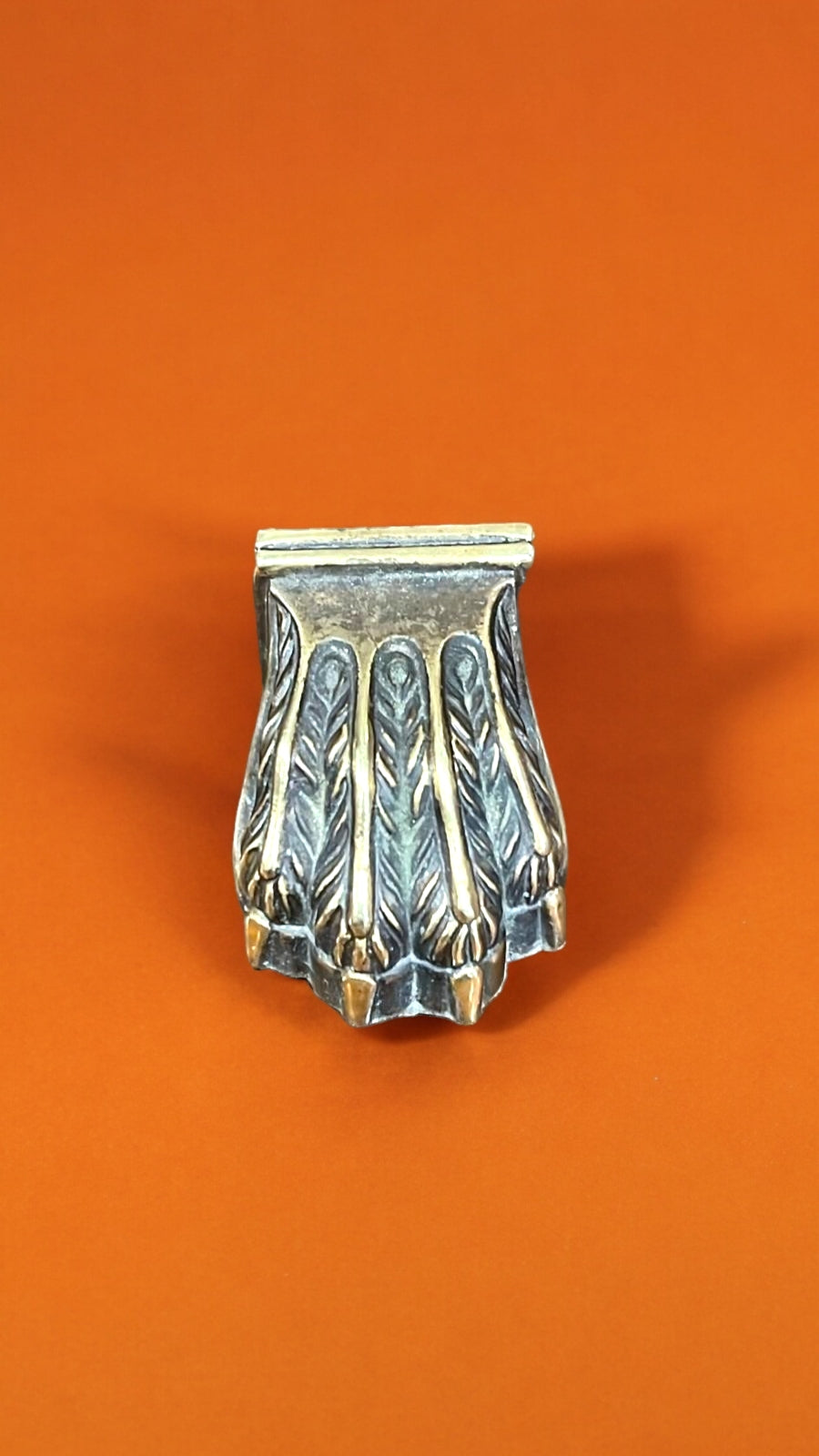 Garra de bronce individual para pata de mesa, con diseño detallado y acabado envejecido, sobre fondo naranja. Ideal para embellecer muebles con estilo.