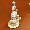 Figura de porcelana Señora con Ovejas vista lateral, muestra detalles florales en el delantal y textura realista en las ovejas. Pieza decorativa refinada.