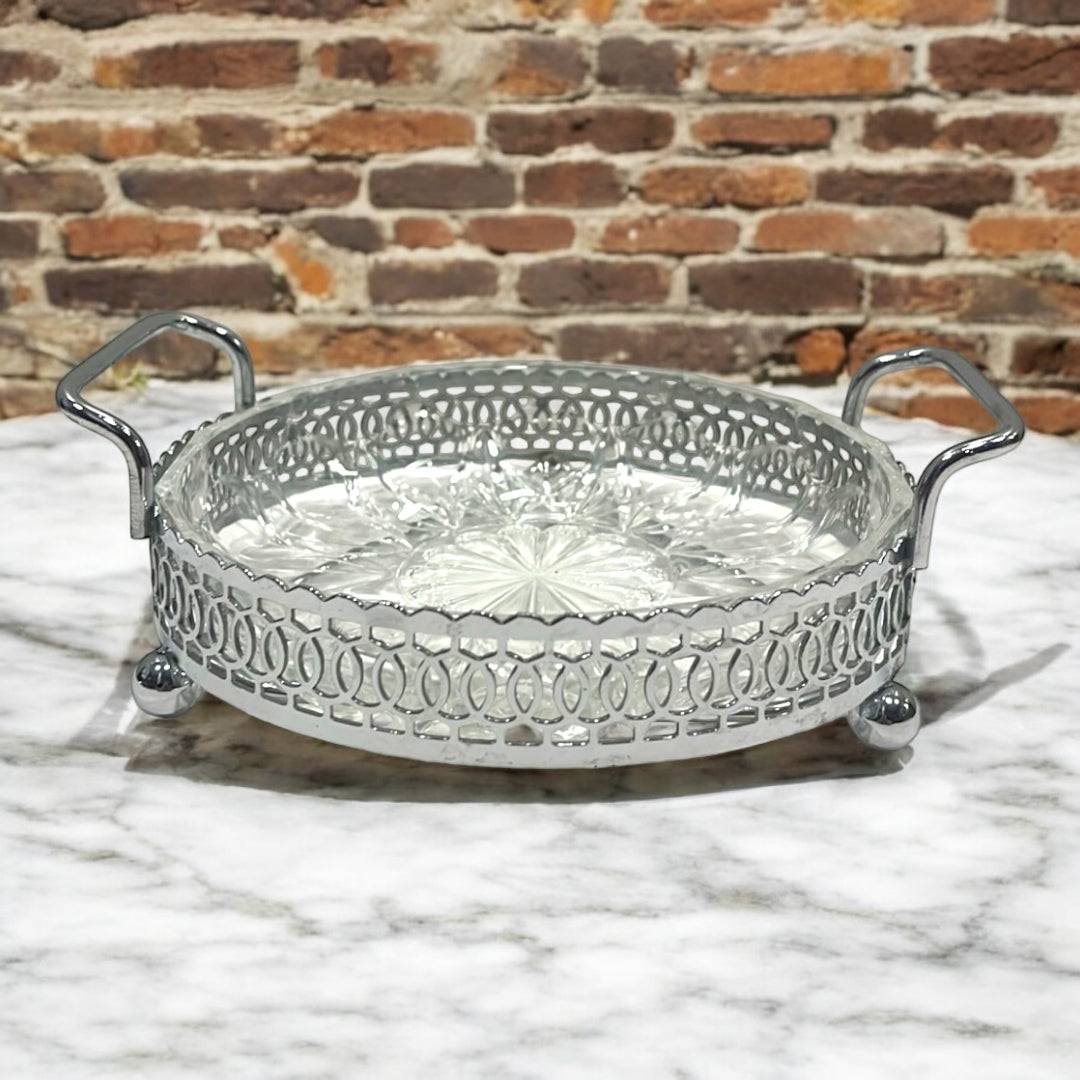 Entremesera de metal plateado y cristal con asas laterales sobre mesa de mármol. Pieza decorativa elegante para el hogar o eventos formales.