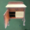Mesita de noche antigua con puerta abierta y compartimentos visibles, incluyendo un espacio con recipiente blanco. Diseño elegante para decoración vintage.