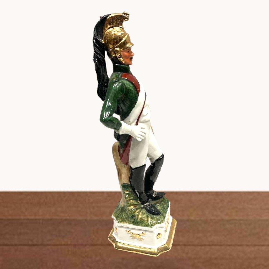 Figura lateral de dragón militar napoleónico en porcelana, con casco dorado y uniforme verde, rojo y blanco sobre base con detalles dorados.