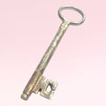 Llave antigua de forja en metal envejecido sobre fondo rosa, ideal como elemento decorativo único para tu hogar en estilo vintage.