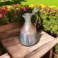 Jarra - Aguamanil antigua de cobre martillado artesanal con tapa abierta, mostrando su interior y detalles forjados a mano en jardín floral.