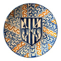 En las imágenes se aprecia una composición muy marcada, centrada en un escudo heráldico en azul sobre fondo claro, rodeado por una decoración radial de bandas geométricas y motivos ondulantes en azul cobalto y ocre anaranjado. 