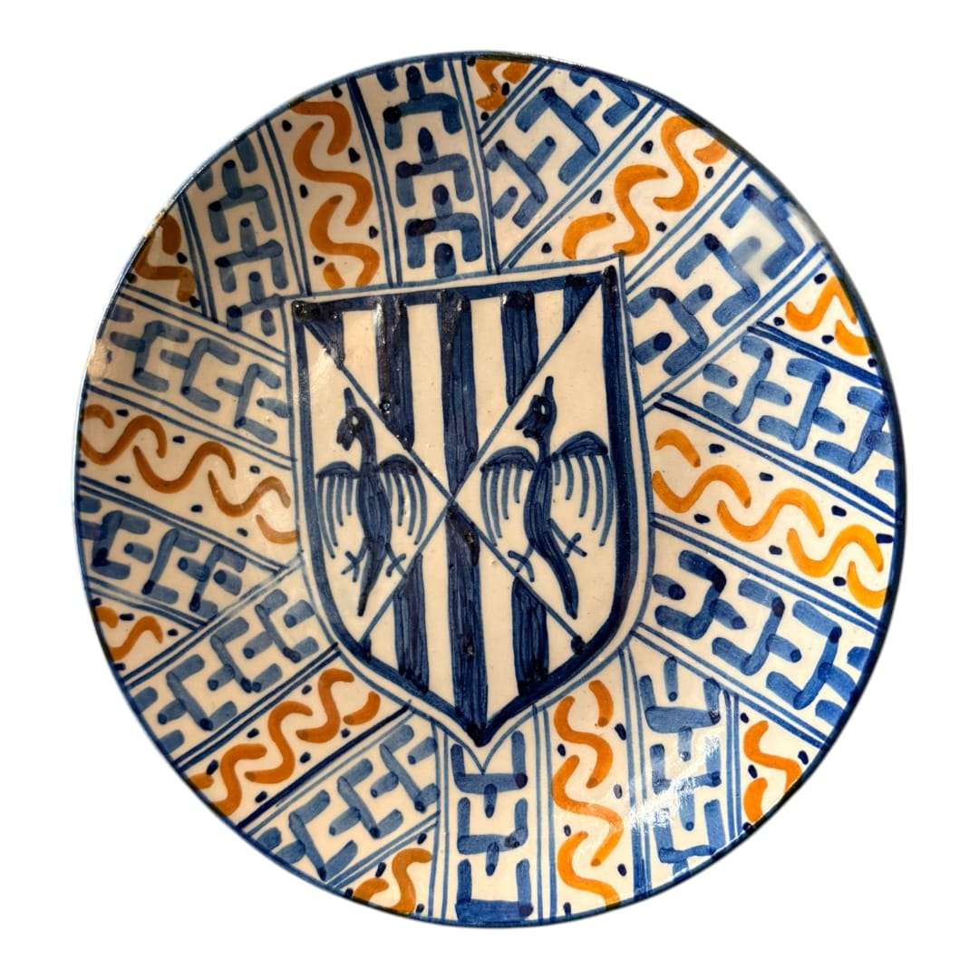 En las imágenes se aprecia una composición muy marcada, centrada en un escudo heráldico en azul sobre fondo claro, rodeado por una decoración radial de bandas geométricas y motivos ondulantes en azul cobalto y ocre anaranjado. 