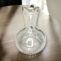 Su diseño transparente permite apreciar el trabajo del cristal, con cuerpo redondeado, cuello estilizado, pico vertedor marcado y asa curva de gran presencia visual. 