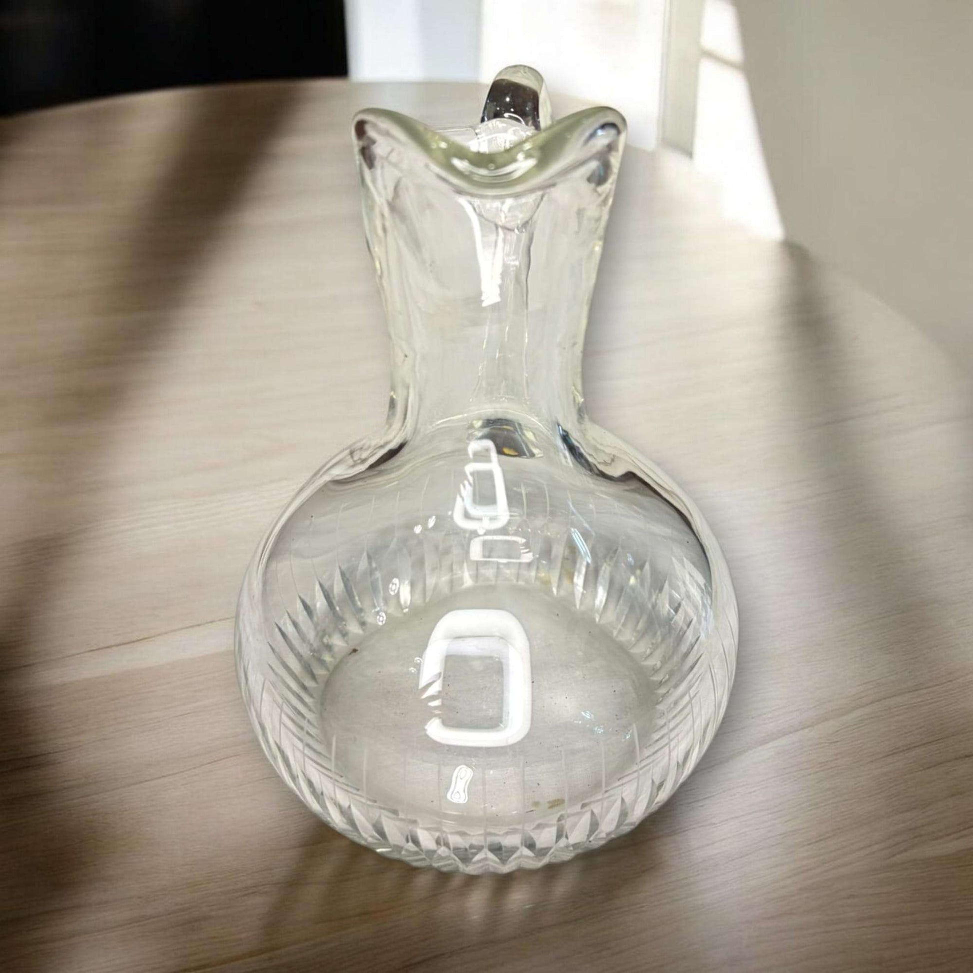 Su diseño transparente permite apreciar el trabajo del cristal, con cuerpo redondeado, cuello estilizado, pico vertedor marcado y asa curva de gran presencia visual. 