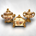 Formado por tres recipientes ornamentales con tapa, combina fondo marfil craquelado, motivos florales multicolor y relieves dorados de gran impacto visual.

Su función original es guardar joyas, pequeños recuerdos, bisutería, llaves, medallas o piezas delicadas. Hoy también destaca como objeto decorativo y de colección, ideal para vitrinas, tocadores, apar