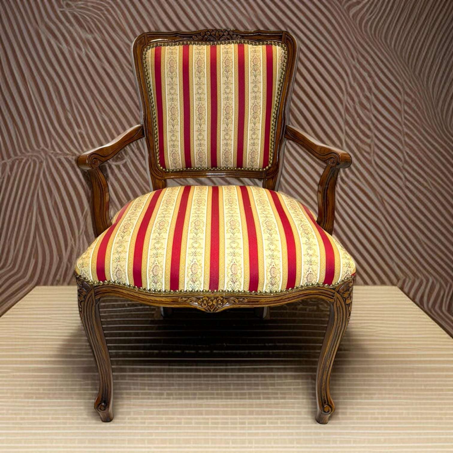 Hoy conserva esa función práctica, pero añade además un gran valor decorativo gracias a su diseño clásico, sus brazos curvados y su tapizado de rayas en tonos rojo, dorado y crema.