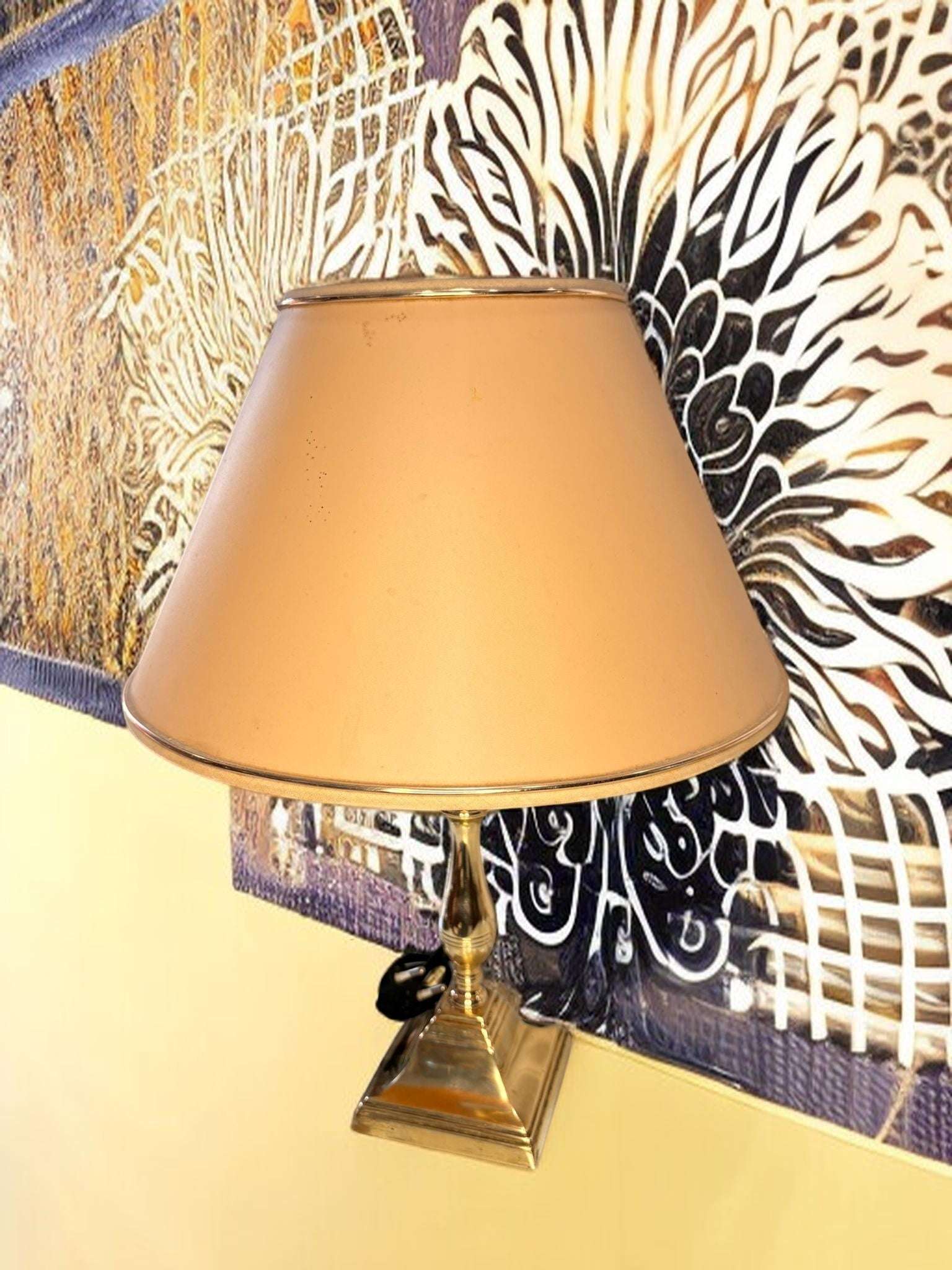 Presenta base cuadrada escalonada de latón, fuste central torneado con formas bulbosas, acabado dorado brillante, pantalla cónica beige con ribete superior e inferior, cable negro visible con interruptor de paso, formato compacto de sobremesa y una estética muy decorativa propia del gusto Hollywood Regency.