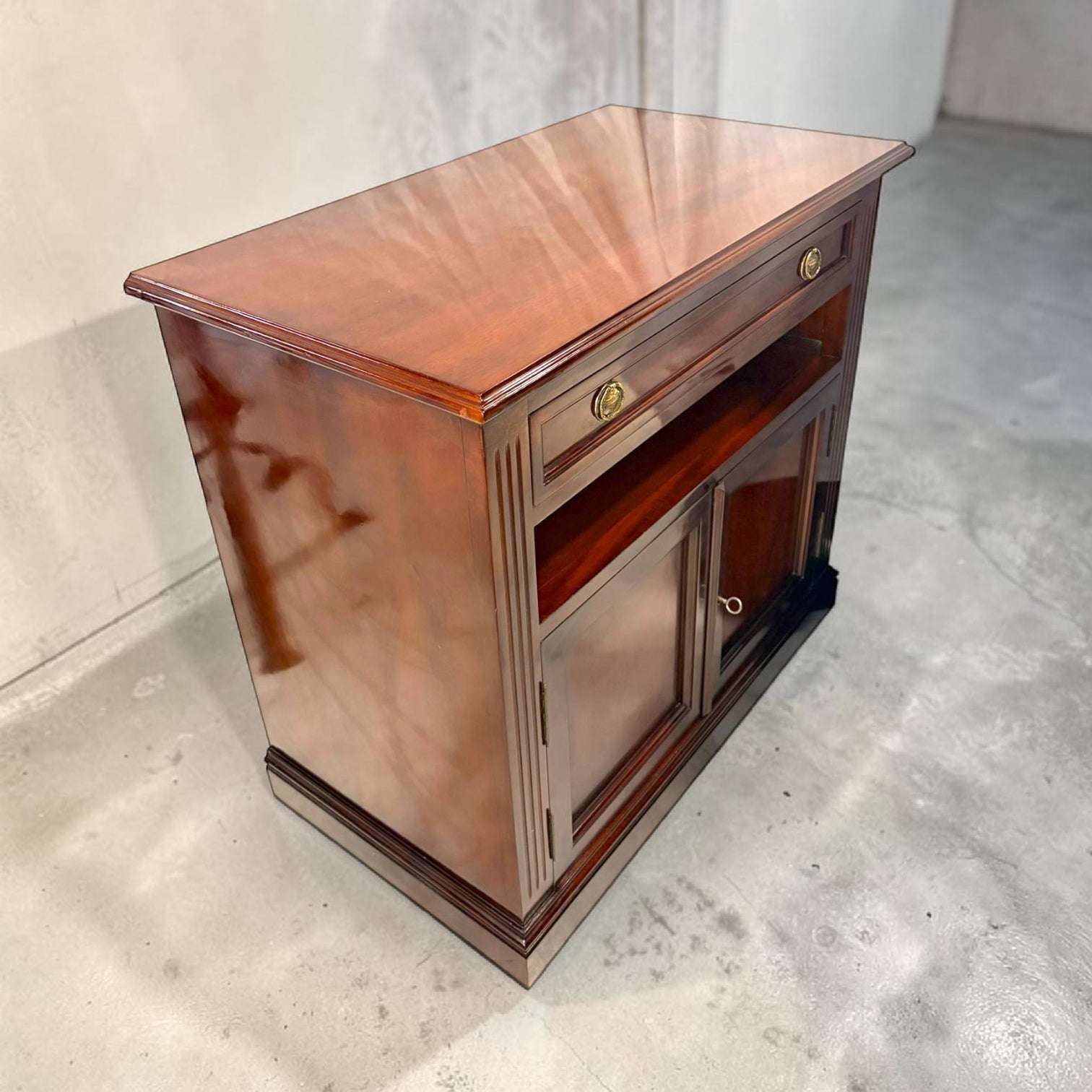 Medidas: 90 x 50 x H82,5 cm. Fabricada en caoba, esta Mesa Tv destaca por su acabado brillante en tono rojizo, su cajón superior con dos tiradores metálicos dorados, su hueco central abierto para aparatos o decoración y su alacena inferior con dos puertas y cerradura con llave.