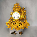 Este Reloj de sobremesa antiguo es una pieza decorativa de gran presencia, realizada en metal dorado y con una composición inspirada en el gusto rococó o Luis XV. 