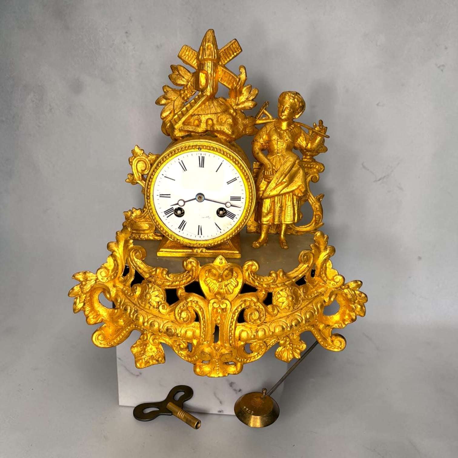 Este Reloj de sobremesa antiguo es una pieza decorativa de gran presencia, realizada en metal dorado y con una composición inspirada en el gusto rococó o Luis XV. 