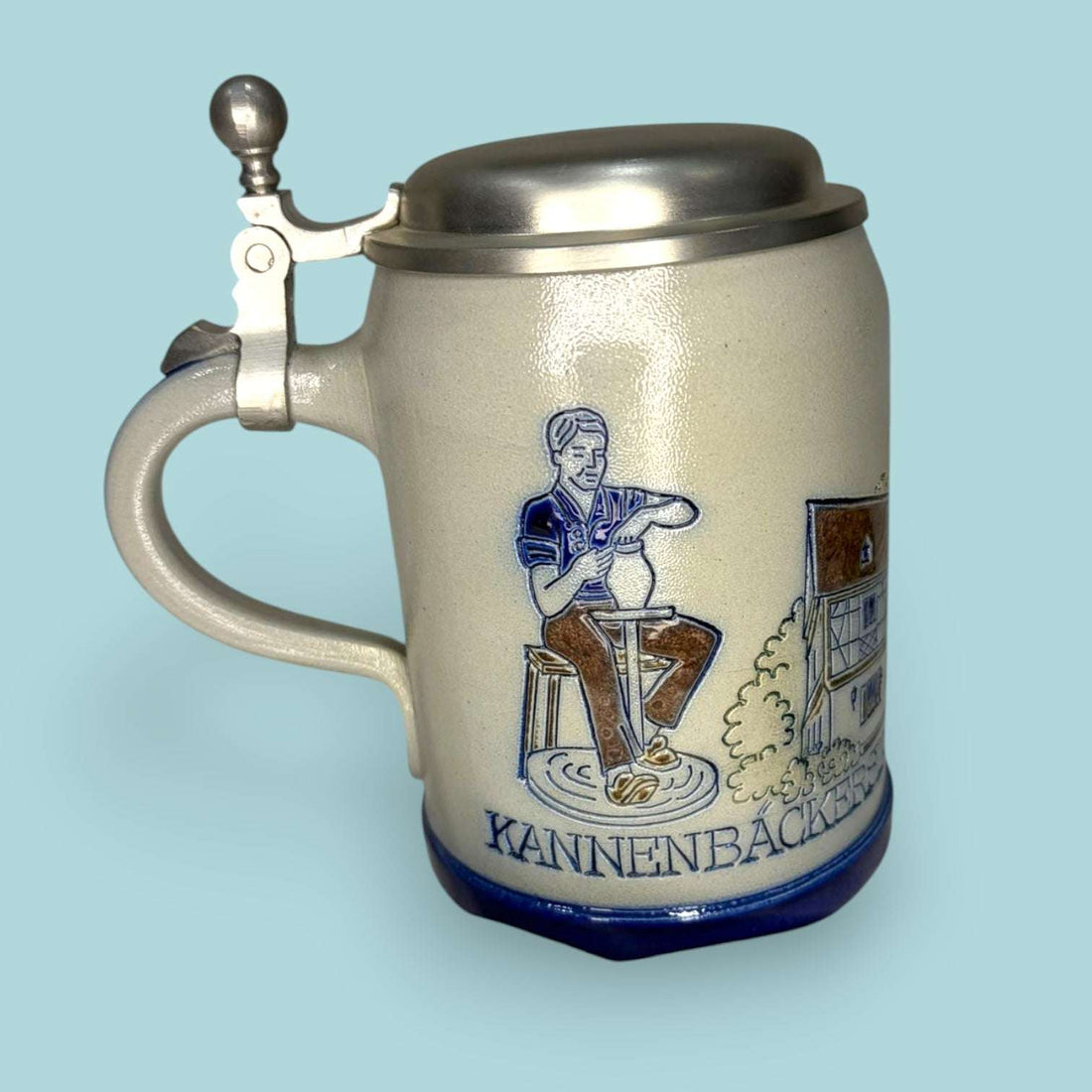 Esta Jarra de cerveza alemana combina cuerpo de cerámica esmaltada, tapa metálica de peltre y una decoración tradicional inspirada en escenas de oficio, arquitectura y paisaje germánico. 