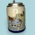 Esta Jarra de cerveza alemana combina cuerpo de cerámica esmaltada, tapa metálica de peltre y una decoración tradicional inspirada en escenas de oficio, arquitectura y paisaje germánico. 