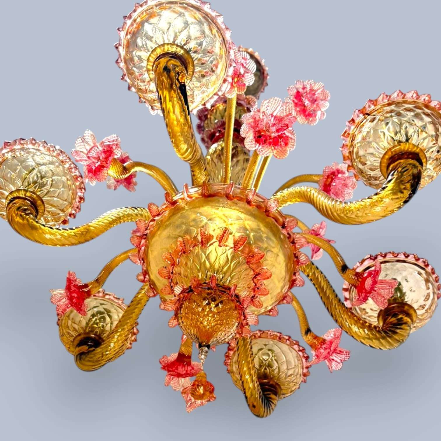 Esta magnífica lámpara destaca por sus delicados detalles florales en tonos rosados, realizados artesanalmente en cristal, combinados con elegantes brazos curvados y tulipas decoradas en tonos ámbar dorado que crean una iluminación cálida y acogedora.