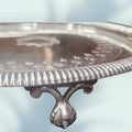Detalle de pata de garra ornamentada en bandeja de metal plateado con decoración floral, perfecta para estilo clásico en decoración del hogar.