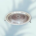 Bandeja de metal plateado con patas de garra y borde acanalado, decoración floral grabada y escudo central, ideal para decoración elegante.