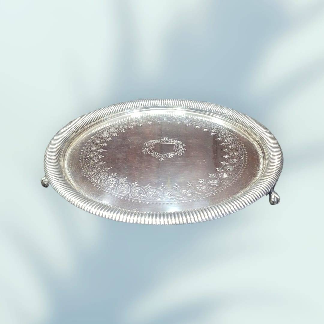 Bandeja de metal plateado con patas de garra y borde acanalado, decoración floral grabada y escudo central, ideal para decoración elegante.
