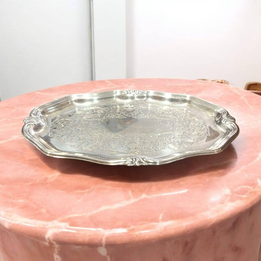 Bandeja ornamental de metal plateado con diseño vintage grabado, colocada sobre mesa de mármol rosa. Pieza decorativa para hogar elegante.