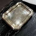 Bandeja de metal plateado con diseño ondulado clásico sobre superficie de mármol negro. Ideal para decoración elegante en el hogar.
