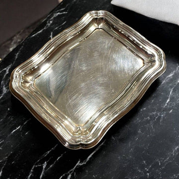 Bandeja de metal plateado con diseño ondulado clásico sobre superficie de mármol negro. Ideal para decoración elegante en el hogar.