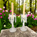 Candeleros de cristal antiguos con detalles colgantes, colocados sobre piedra en jardín, reflejan elegancia y tradición decorativa.