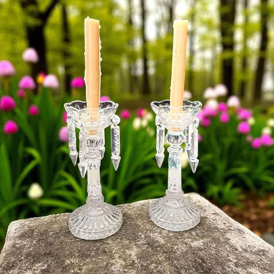 Candeleros de cristal antiguos con detalles colgantes, colocados sobre piedra en jardín, reflejan elegancia y tradición decorativa.