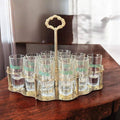 Cañero vintage de metal dorado con 12 vasos de cristal Juncal sobre mesa de madera. Ideal para decoración retro en hogar o bar.