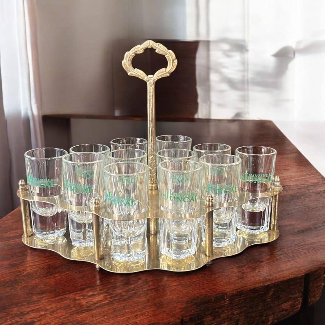 Cañero vintage de metal dorado con 12 vasos de cristal Juncal sobre mesa de madera. Ideal para decoración retro en hogar o bar.