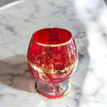 Copa de cristal Murano rojo con detalles dorados sobre mesa de mármol. Diseño artesanal que combina lujo y sofisticación en cada detalle.