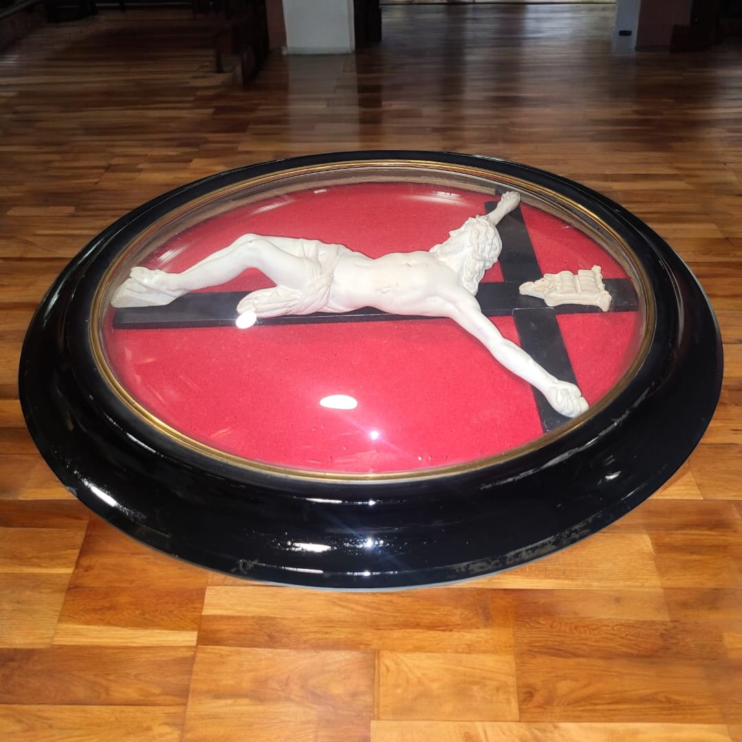 Vista inclinada del crucifijo, donde se aprecia la profundidad de la cúpula de cristal y el contraste entre el fondo rojo y la escultura blanca. Una pieza única de arte sacro.