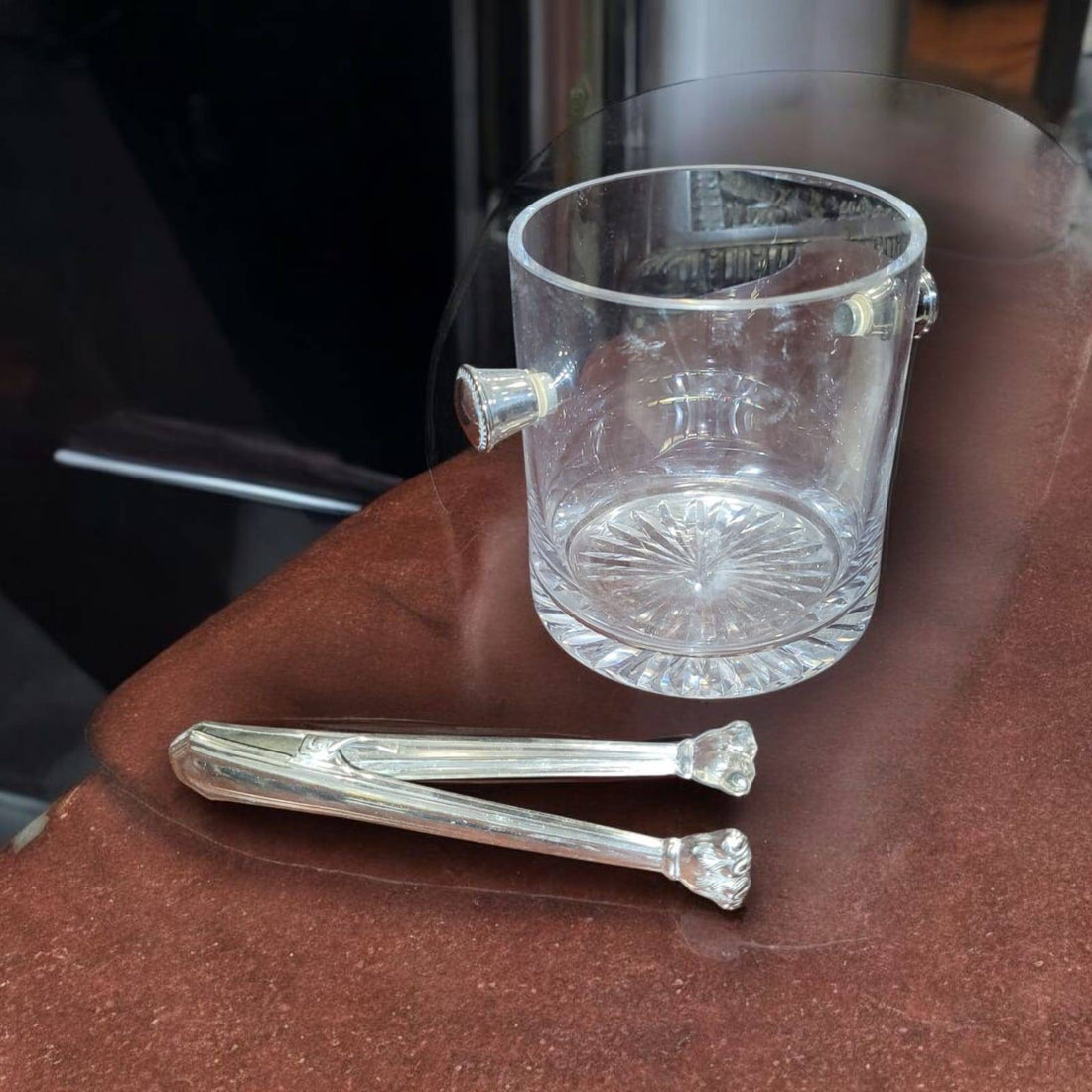 Cubitera de cristal tallado con asas laterales y pinzas de metal plateado sobre mesa marrón. Ideal para decorar y servir hielo con estilo.