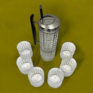 Coctelera de cristal con tapa de plata y seis vasos a juego sobre fondo verde, ideal para decoración elegante en eventos en casa.