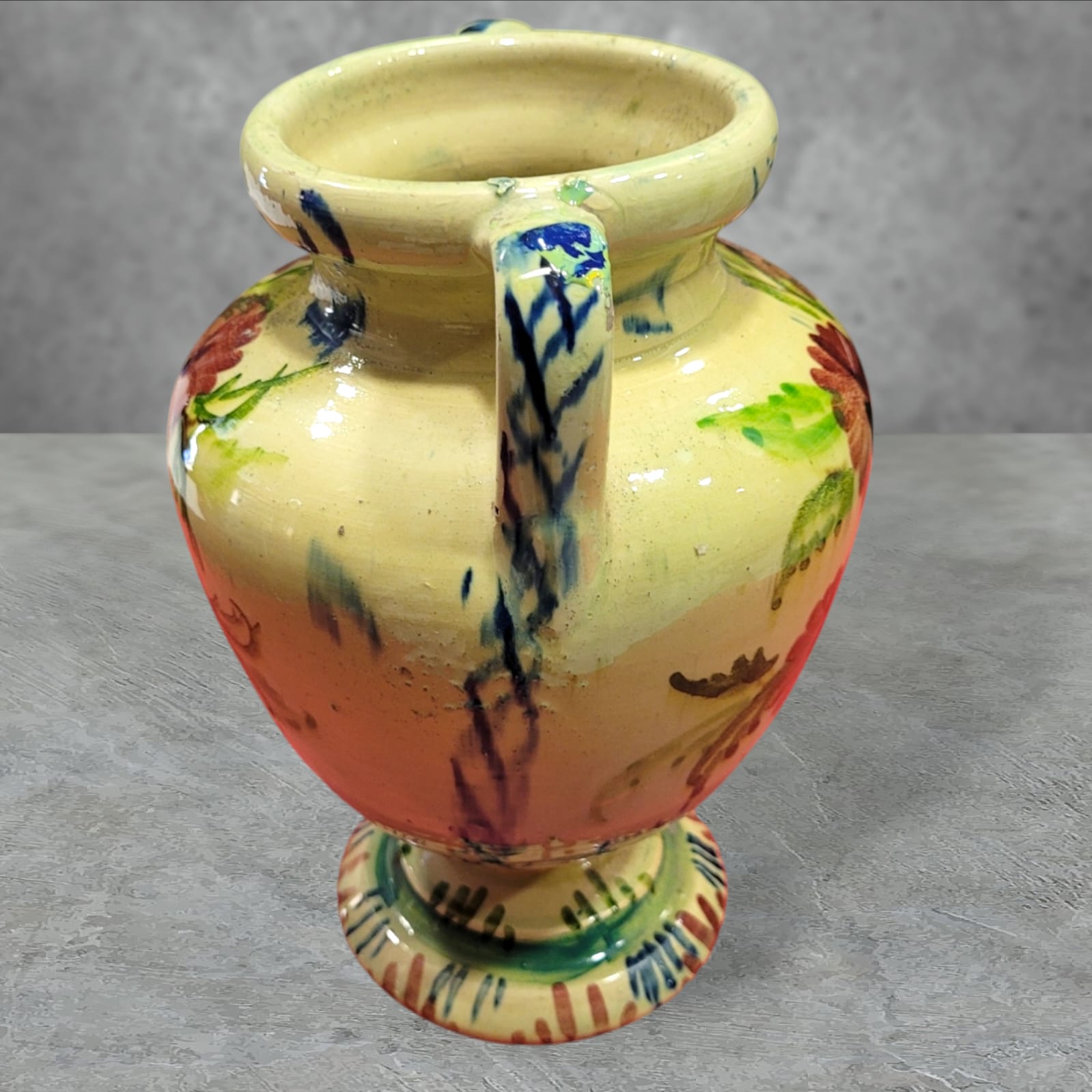 Vista lateral de ánfora de cerámica esmaltada con asas y detalles florales pintados a mano. Perfecta para añadir un toque artístico a cualquier espacio.