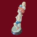 Figura decorativa niño con globos estilo Capodimonte vista en ángulo, con ropa veraniega y base de madera. Pieza única para colección.