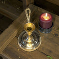 Palmatoria antigua de metal dorado con vela encendida al lado, destacando su mango en relieve y estilo decorativo clásico.