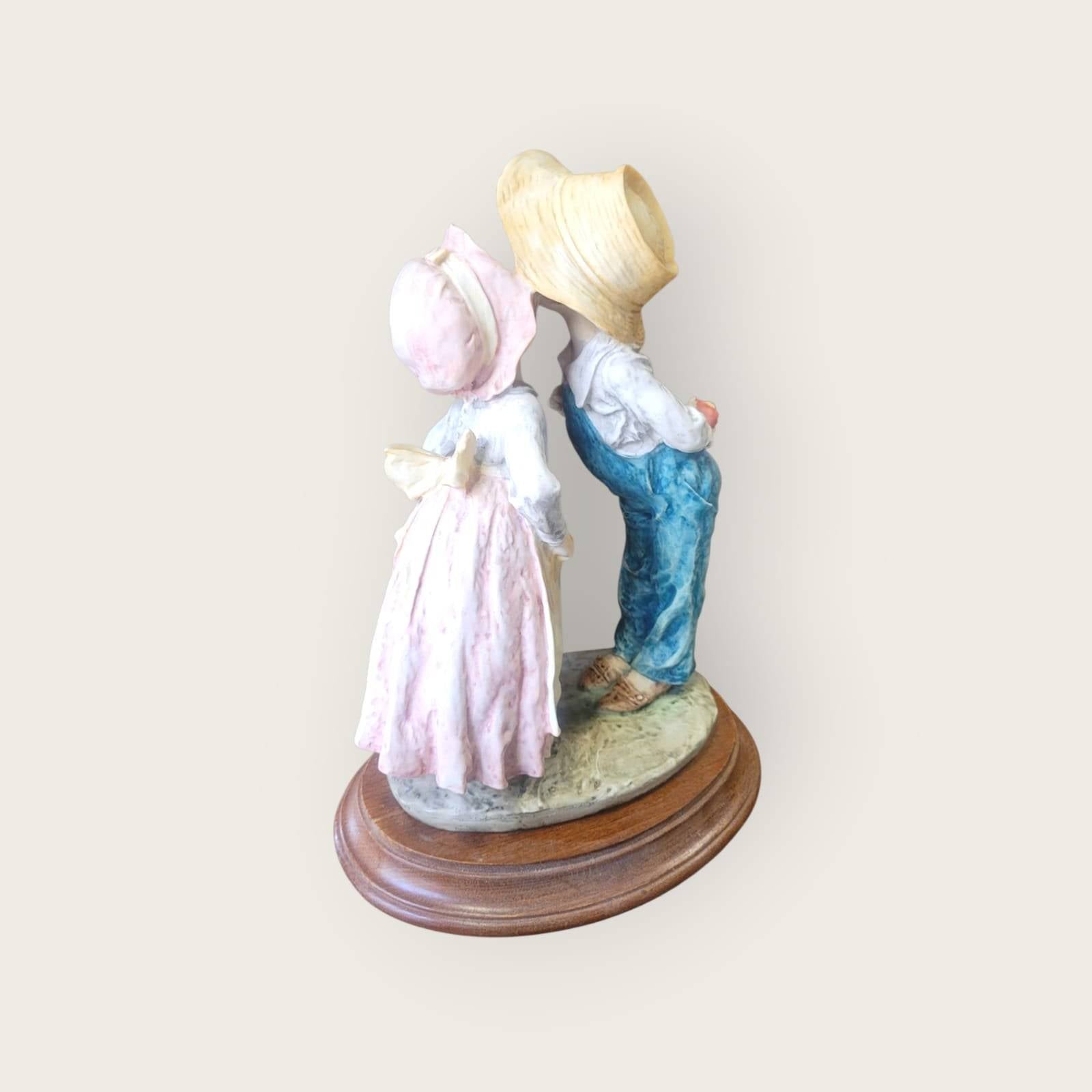 Figura de pareja infantil en resina sobre base de madera, mostrando un beso tímido desde ángulo posterior, perfecta para colecciones románticas.