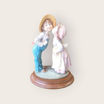 Figura Beso Tímido Resina - Romántica sobre base de madera, muestra a una pareja infantil en gesto de beso, ideal para decoración del hogar.