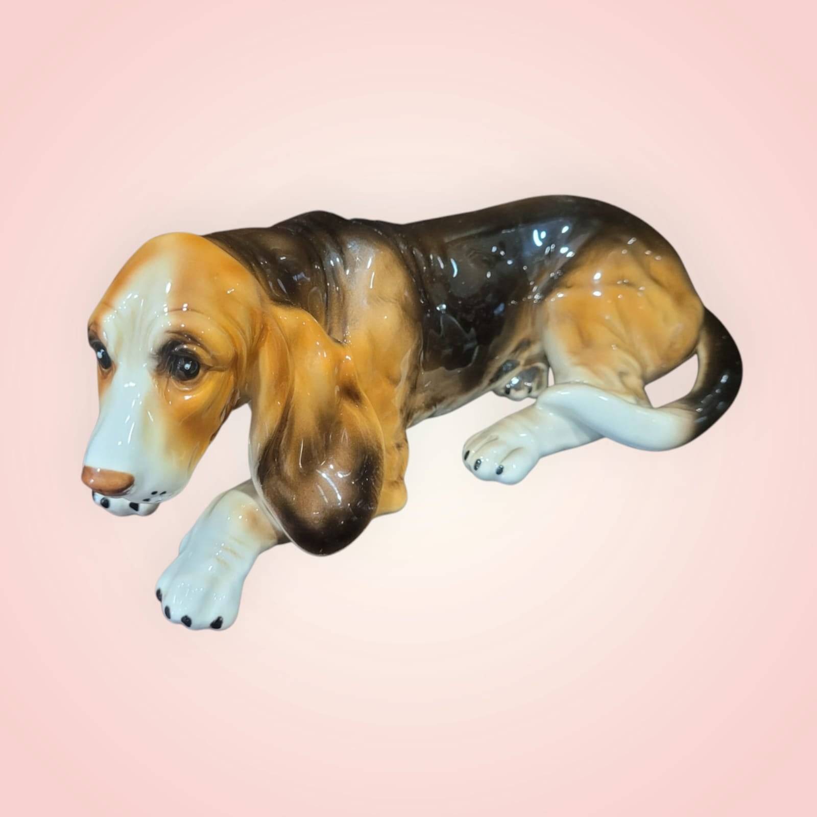 Perro de porcelana Basset Hound en posición recostada, con expresión tierna y esmaltado detallado. Figura decorativa para amantes de los perros.